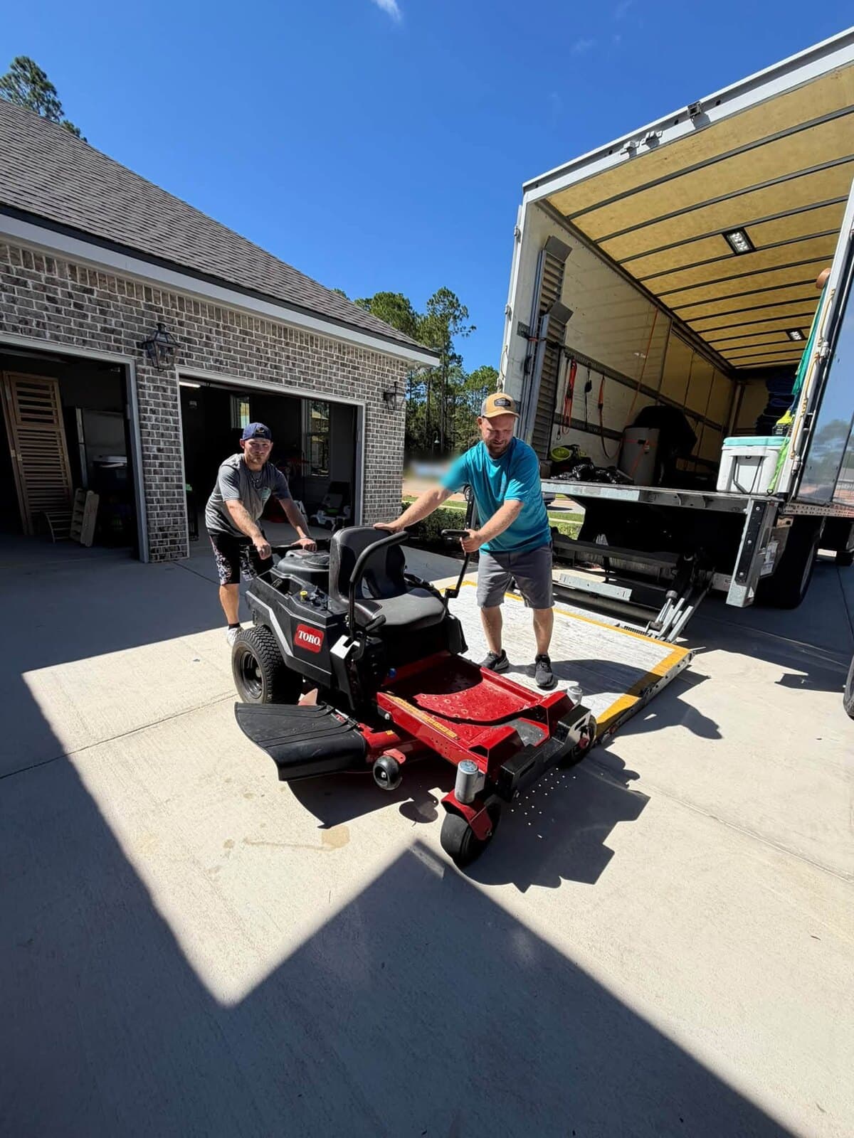 Two Beach House Moving movers rolling a red zero-turn mower from a garage toward the box truck lift gate in Hammock Bay, Freeport, FL