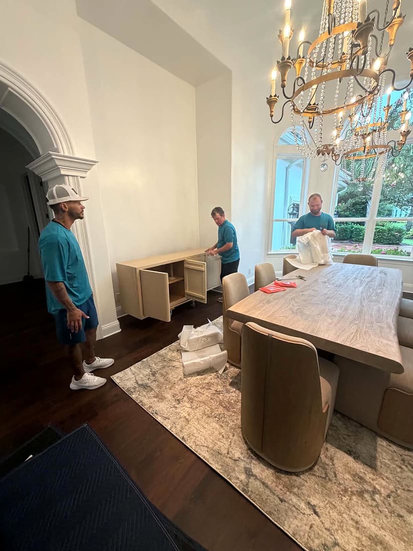 Beach House Moving crew unwrapping and placing dining room furniture under a chandelier at a Burnt Pine home in Miramar Beach, FL