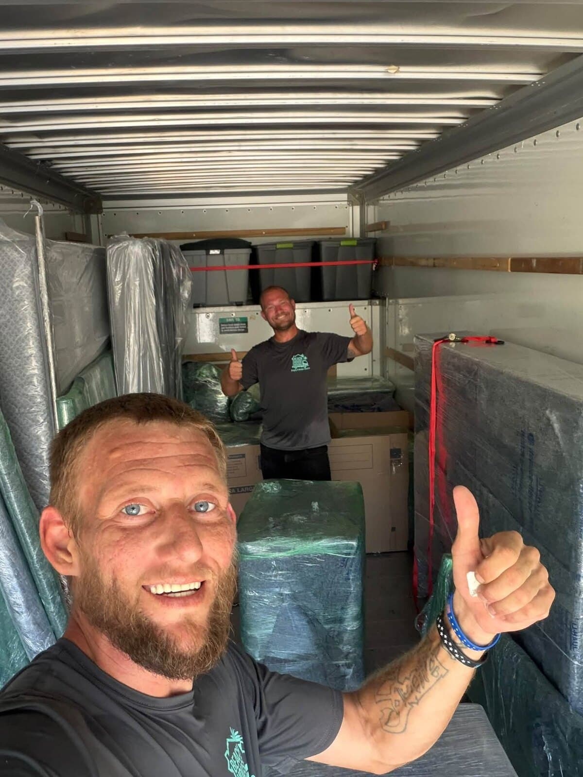 Two Beach House Moving crew members giving a thumbs-up inside a truck loaded with wrapped furniture and strapped bins
