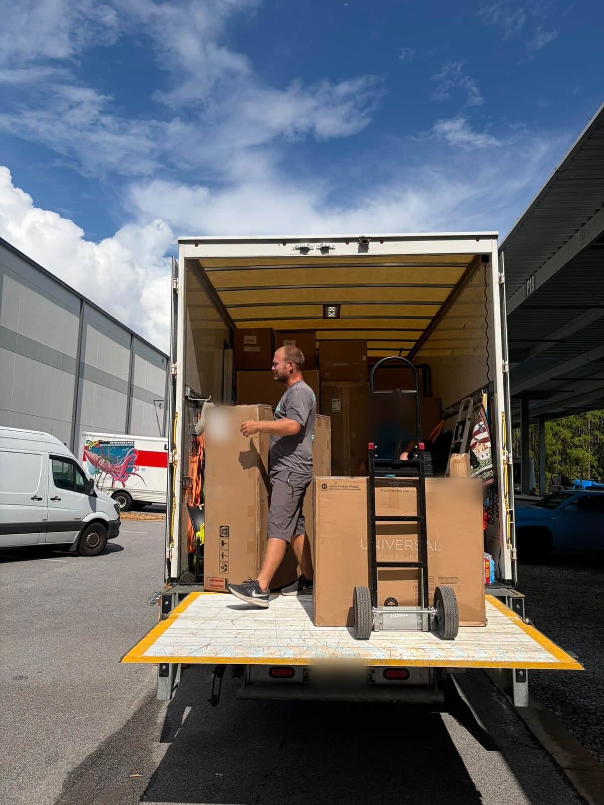 Beach House Moving crew loading boxed furniture onto a box truck lift gate at a storage facility the day before a design install