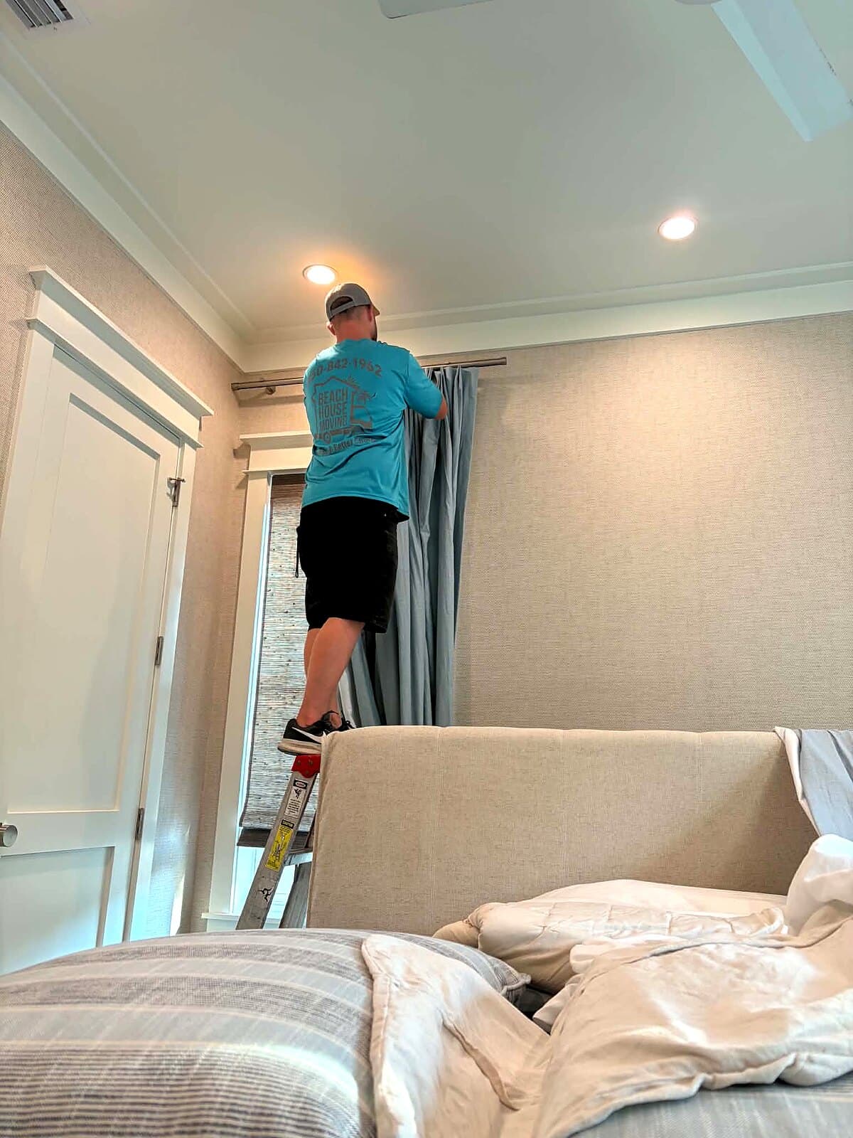 Beach House Moving mover Zack on a ladder hanging drapery panels in a Santa Rosa Beach, FL primary bedroom